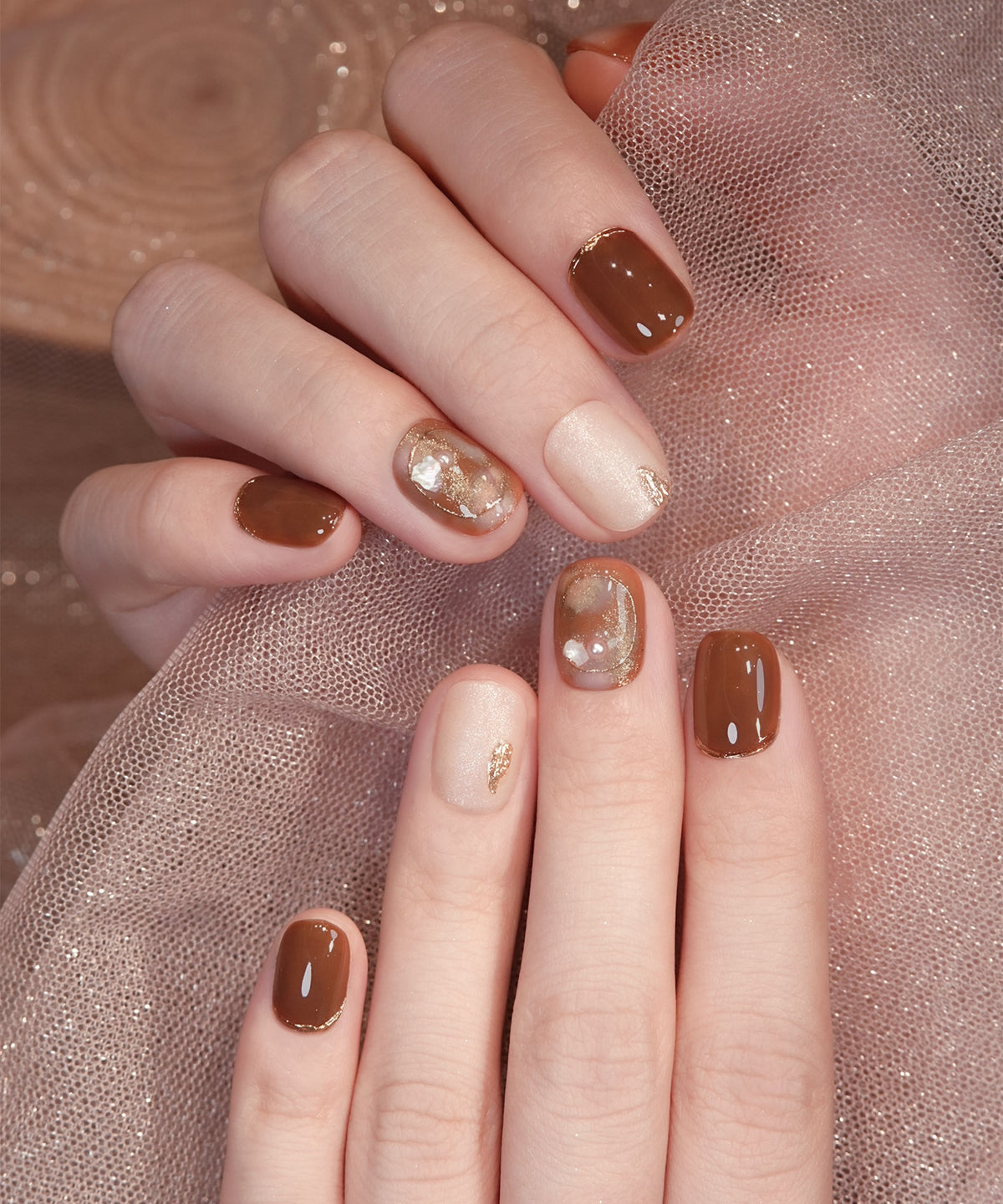 深みのあるブラウンにシェルやパール、マットラメとゴールドミラーアートを施した、ショートラウンド型のジェルネイルチップを手元に着けた様子。